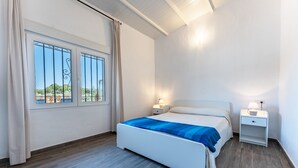 Une chambre à coucher avec un lit, deux tables de chevet, une fenêtre avec des rideaux et une vue sur la verdure à l’extérieur.