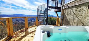 Un jacuzzi en una terraza de madera con vistas a la ciudad, una escalera de caracol y una pared de piedra.