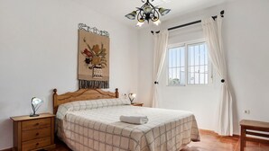 Een slaapkamer met een houten bed, een nachtkastje met een lamp, een kunstwerk aan de muur, een raam met gordijnen en een houten bankje.