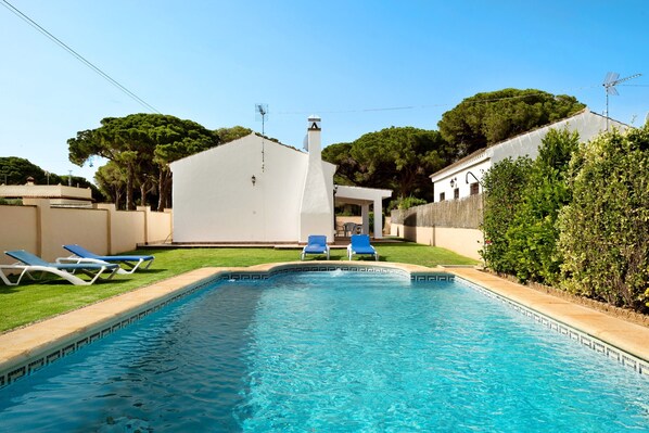 A swimming pool with lounge chairs, a white building, and trees in the background.