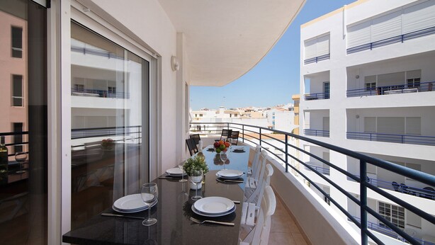 A balcony with a dining table set for two, a potted plant, and a view of neighboring buildings. - Apartment 'Quarteira Beach Iii' with Balcony, Wi-Fi and Air Conditioning (Quarteira)