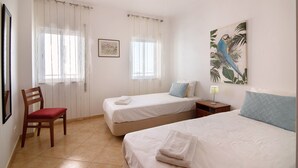 A room with two beds, a chair, a nightstand, and a painting. - Apartment 'Quarteira Beach Iii' with Balcony, Wi-Fi and Air Conditioning (Quarteira)