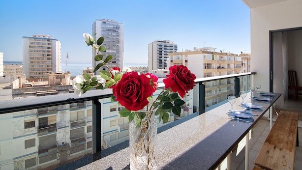 A balcony with a vase of red roses, a dining table set with plates and glasses, and a view of city buildings and the sea. - Apartment 'Quarteira Beach I' with Balcony, Wi-Fi and Air Conditioning (Quarteira)