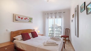 A bedroom with a bed, a nightstand, a chair, a window with curtains, and a painting on the wall. - Apartment 'Quarteira Beach I' with Balcony, Wi-Fi and Air Conditioning (Quarteira)