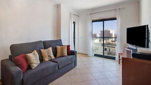 A living room with a sofa, a television, and a view through a large window. - Apartment 'Quarteira Beach I' with Balcony, Wi-Fi and Air Conditioning (Quarteira)