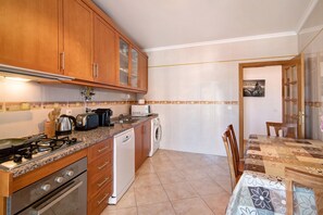 Une cuisine avec des meubles de rangement en bois, une cuisinière à gaz, un lave-vaisselle et une table recouverte d’une nappe à motifs floraux.