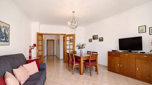 A living room with a sofa, a dining table with chairs, a TV, and a wooden cabinet. - Apartment 'Quarteira Beach Ii' with Balcony, Wi-Fi and Air Conditioning (Quarteira)