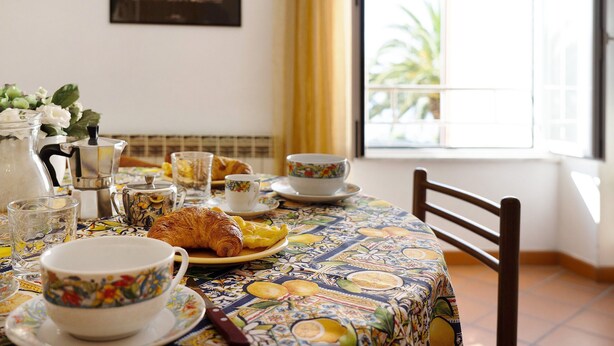 Una mesa de desayuno con mantel estampado, un croissant, una cafetera, tazas y una ventana con cortinas.