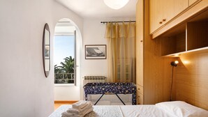 Un dormitorio con una cama, una mesita con toallas, una ventana con cortinas y un cuadro en la pared.
