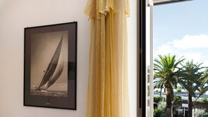 Fotografía enmarco de un velero en una pared, una ventana con vista a palmeras y un edificio, y una cortina.