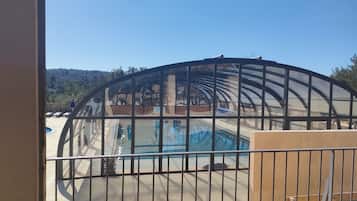 Une piscine extérieure couverte, dotée d’une structure en verre, d’une terrasse en bois et offrant une vue sur un paysage vallonné.
