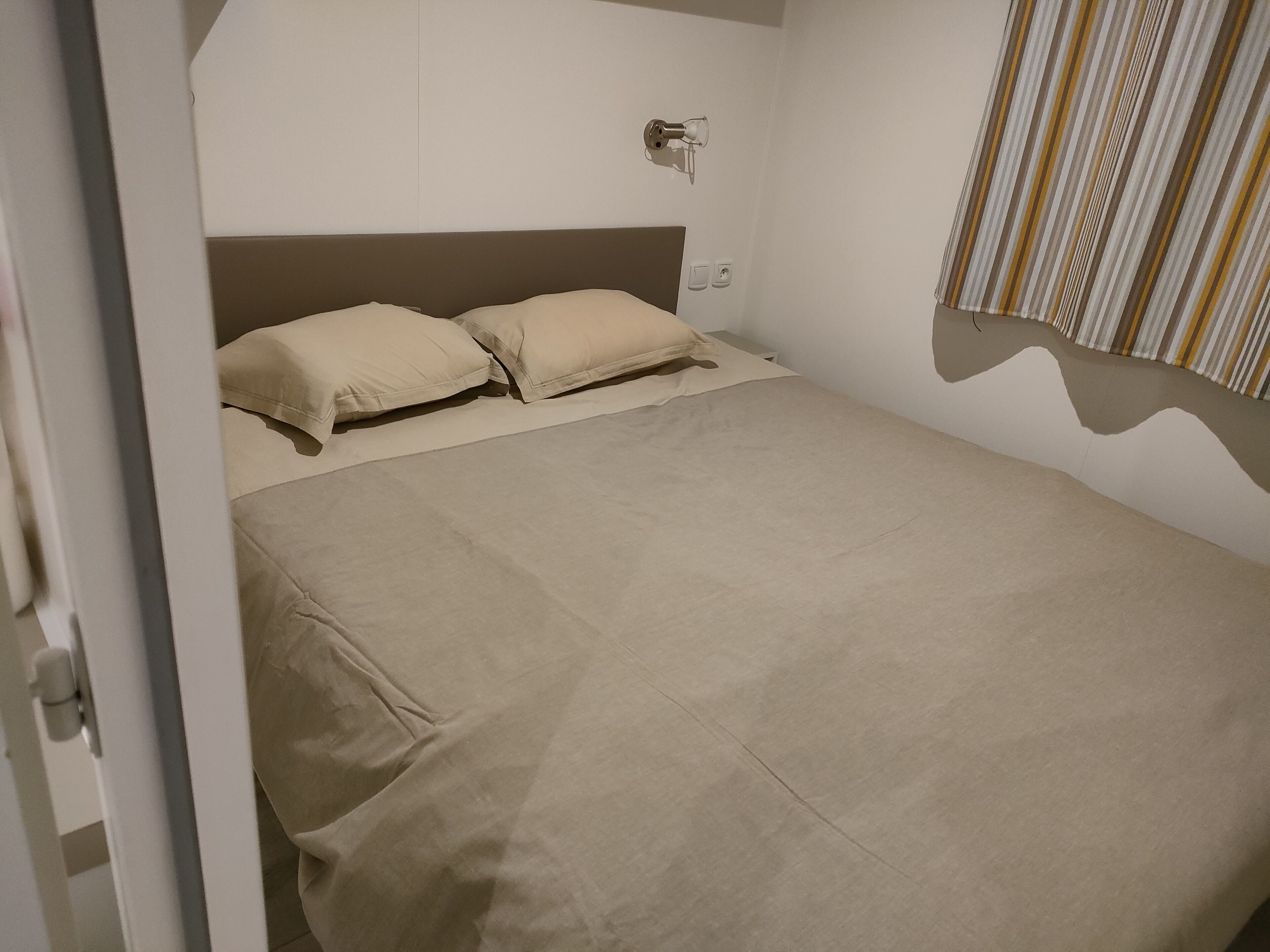 Un lit bien fait, avec une literie beige, dans une pièce aux murs blancs et ornée d’un rideau à rayures.