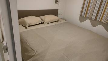 Un lit bien fait, avec une literie beige, dans une pièce aux murs blancs et ornée d’un rideau à rayures.