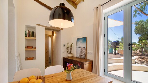 A dining area with a wooden table, a bowl of oranges, a vase with flowers, a pendant light, a bookshelf, and a sliding glass door leading to an outdoor area. - Holiday Home Casa Burro with Mountain View, Shared Pool, Garden & Wi-Fi (Moncarapacho)
