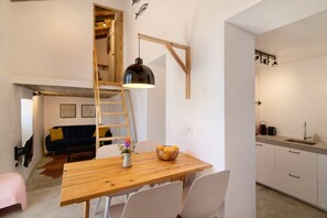 Un espace de vie compact comprenant une table à manger en bois, des chaises blanches, une chambre en mezzanine accessible par une échelle en bois, et un coin cuisine avec un évier et un plan de travail.