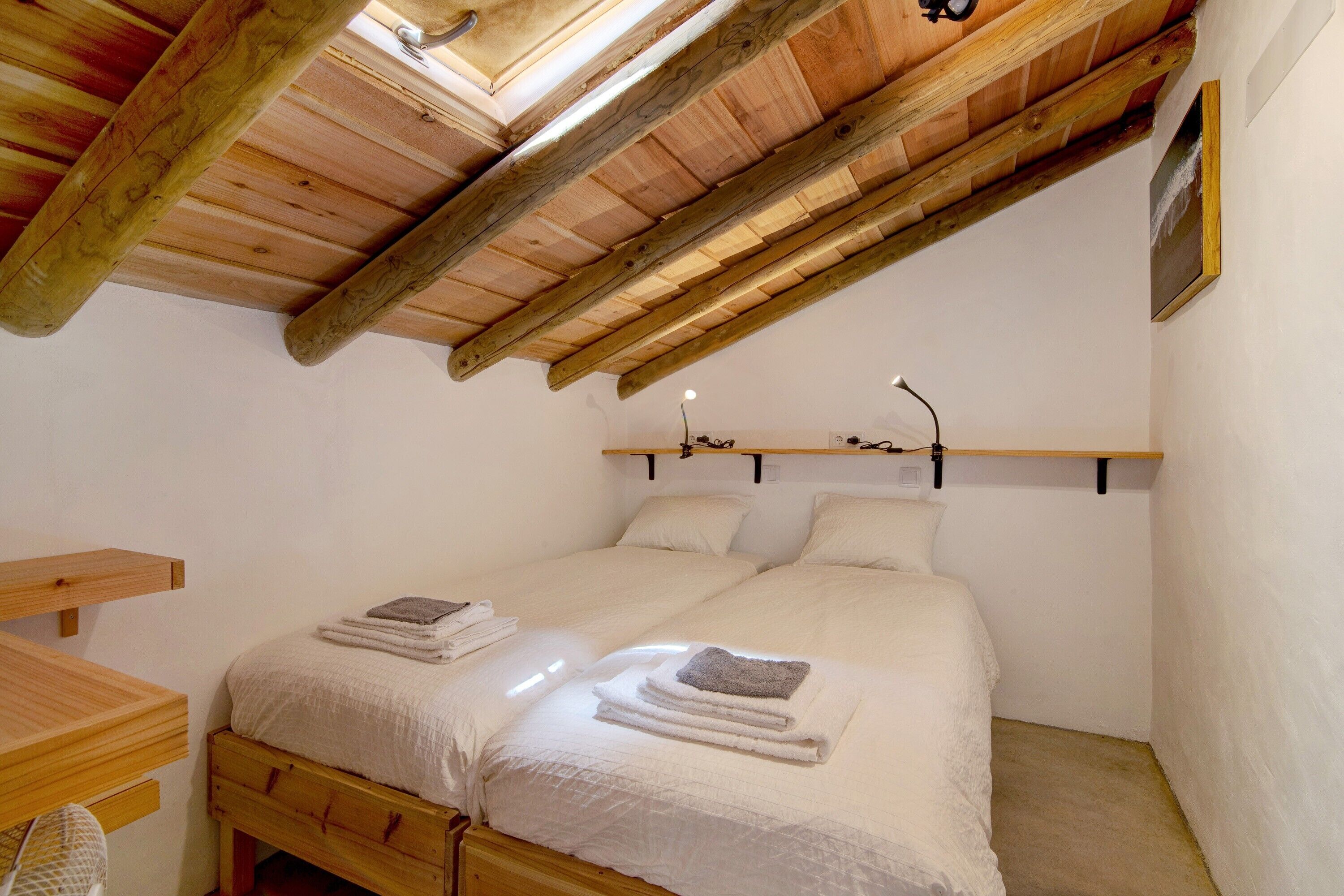 Une chambre avec un plafond en bois, deux lits, une table de chevet et une étagère fixée au mur.
