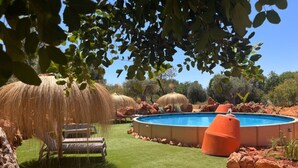 A swimming pool surrounded by rocks and grass, with a thatched umbrella and a wooden chair. - Holiday Home Casa Burro with Mountain View, Shared Pool, Garden & Wi-Fi (Moncarapacho)