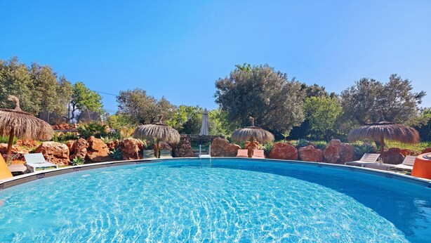 A swimming pool with clear water, surrounded by rocks and thatched umbrellas, and trees in the background. - Holiday Home 'Casa Osga' with Mountain View, Shared Pool, Terrace & Wi-Fi (Moncarapacho)
