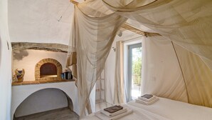 A bedroom with a stone fireplace, a bed with a white bedspread, and sheer curtains. - Holiday Home 'Casa Osga' with Mountain View, Shared Pool, Terrace & Wi-Fi (Moncarapacho)
