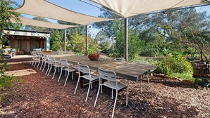 An outdoor dining area with a long table and chairs, shaded by a large canopy, surrounded by mulch and greenery. - Holiday Home 'Casa Osga' with Mountain View, Shared Pool, Terrace & Wi-Fi (Moncarapacho)