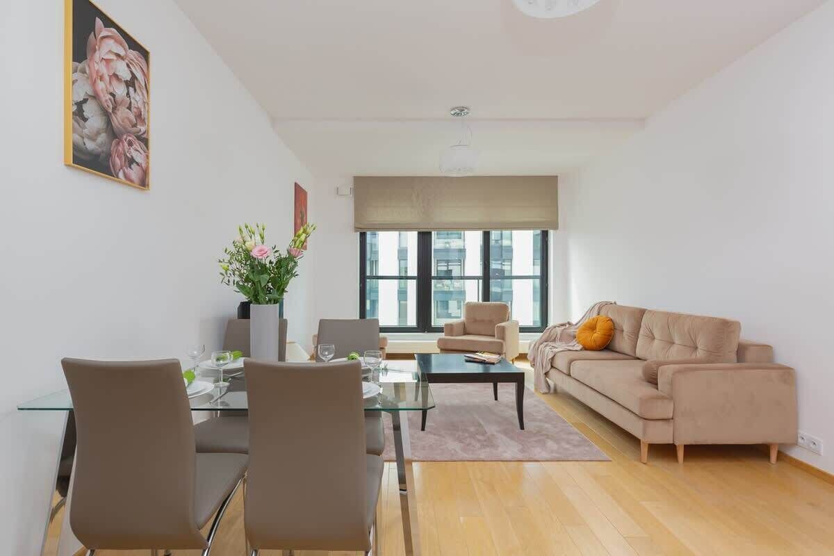 Una sala de estar moderna con una mesa de comedor de vidrio, sofás beige y un gran ventanal con persianas.