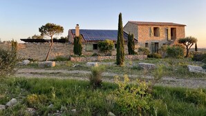 Una casa de piedra con techo de tejas y paneles solares, rodeada por un paisaje natural con árboles y rocas.