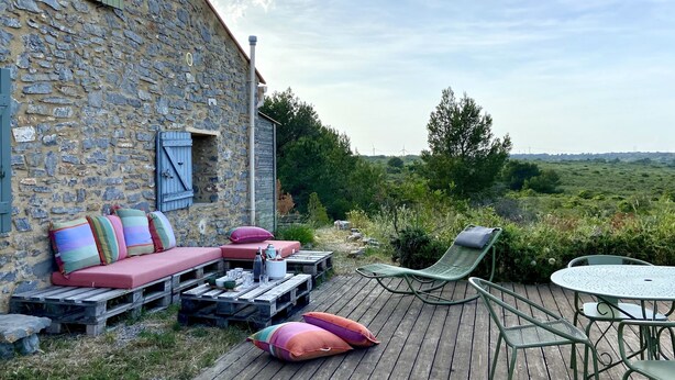 Un edificio rústico de piedra con una terraza de madera, asientos al aire libre y una mesa.