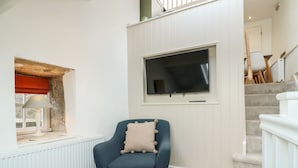 A living room with a blue armchair, a television mounted on the wall, a window with a red blind, and a staircase in the background. - The Hayloft (Matlock)