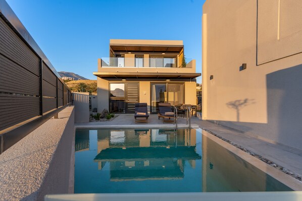 A modern two-story house with a swimming pool, outdoor seating, and a view of the hills.