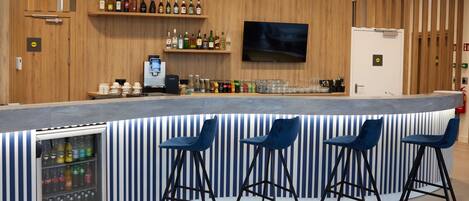Zona de bar con barra, taburetes, refrigerador con bebidas, estanterías con botellas y un televisor montado en la pared.