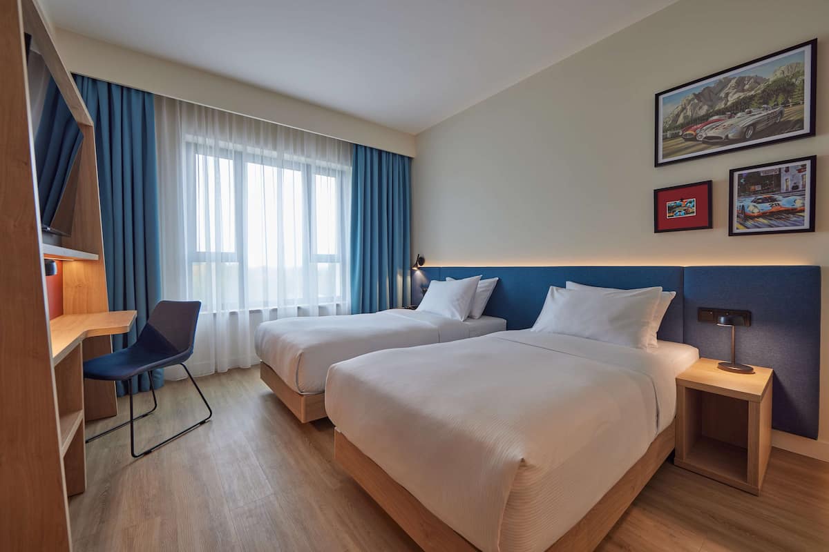 A hotel room with two beds, a desk, a chair, and a nightstand with a lamp.