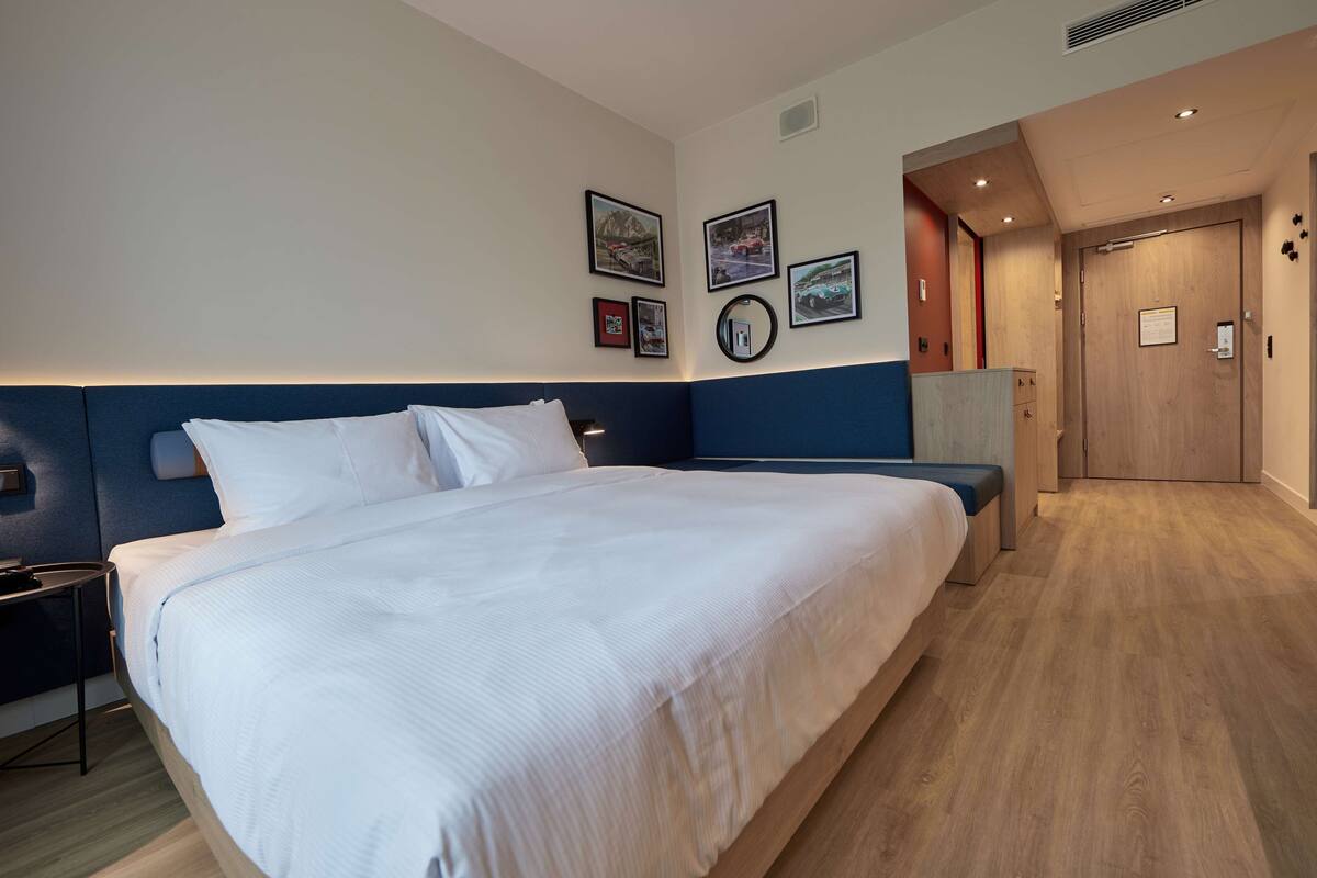 A modern hotel room with a large bed, a bedside table, a wardrobe, and a door leading to another room.