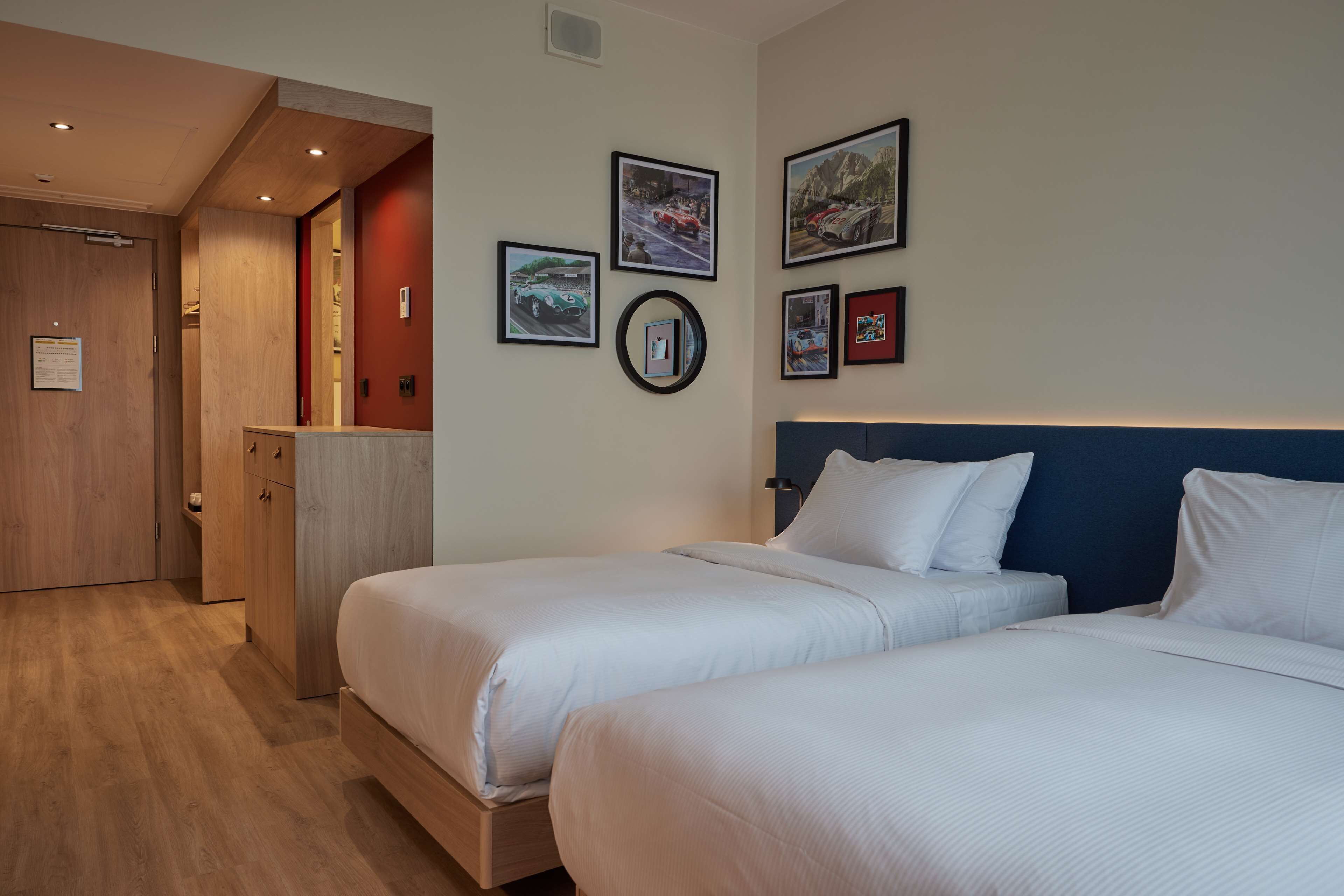A hotel room with two beds, wooden flooring, a wooden wardrobe, and a wall with framed pictures.