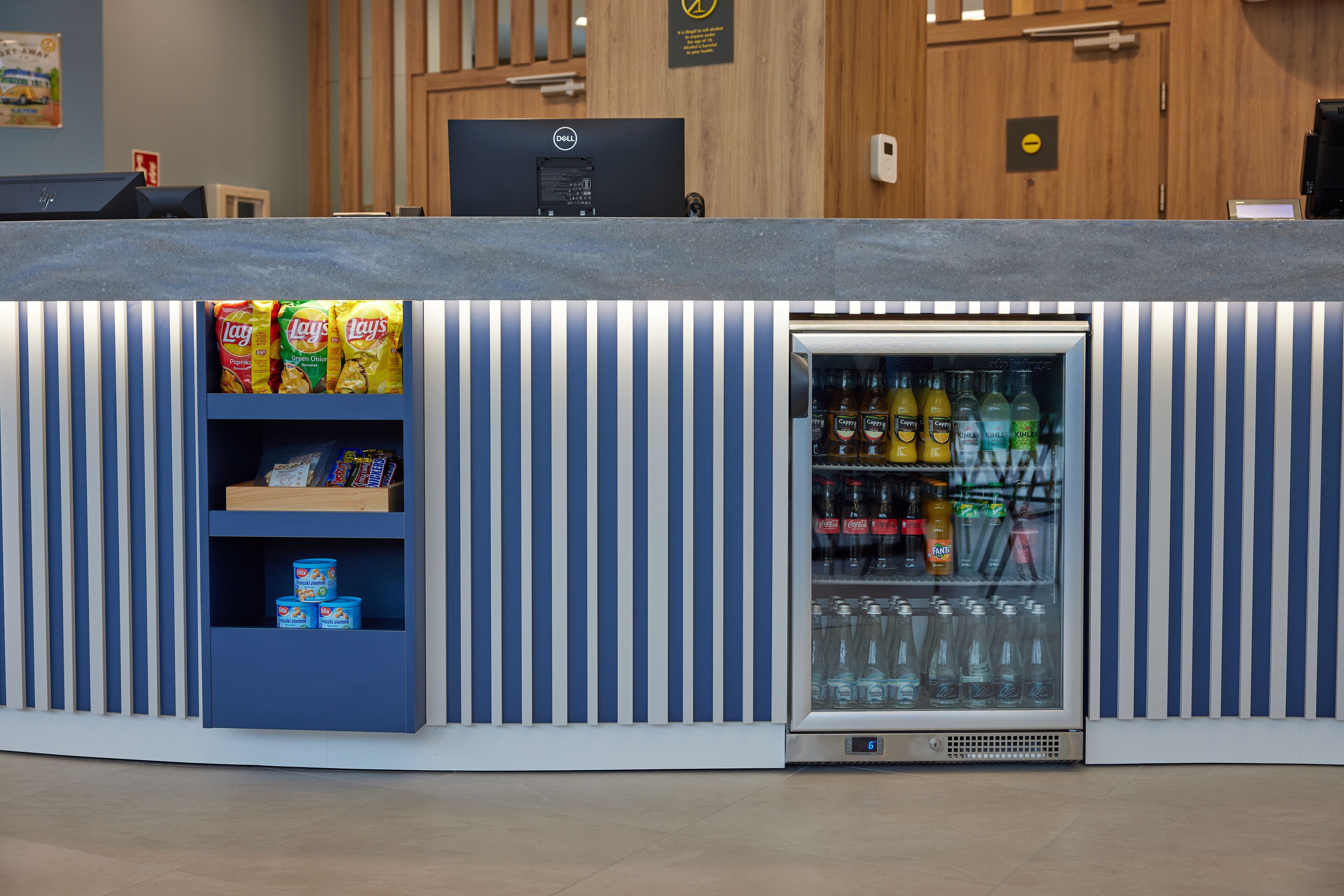 Zona de recepción con una computadora Dell, estantes con aperitivos y un refrigerador para bebidas.