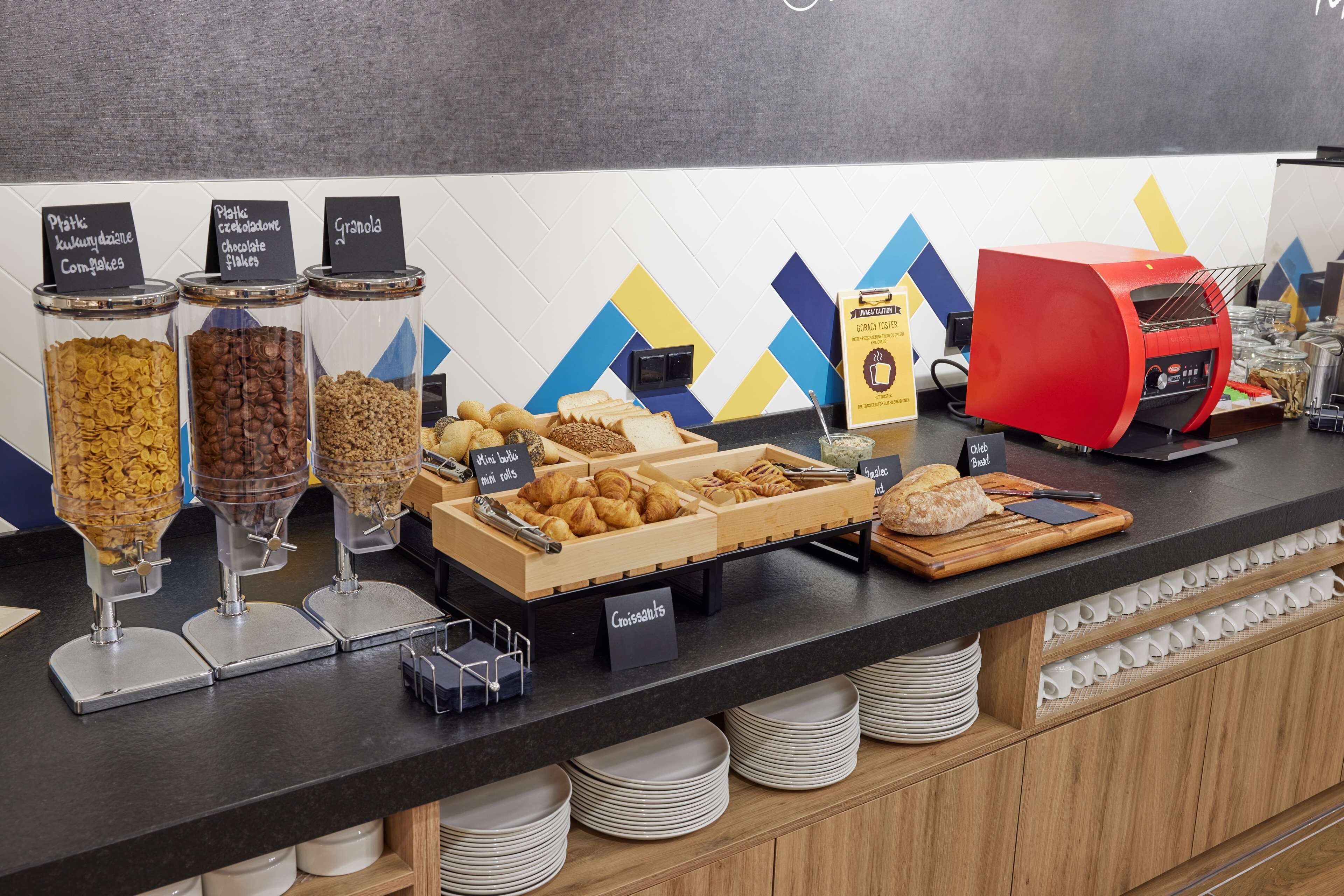 A breakfast buffet with cereal, chocolate flakes, granola, mini rolls, croissants, and bread.