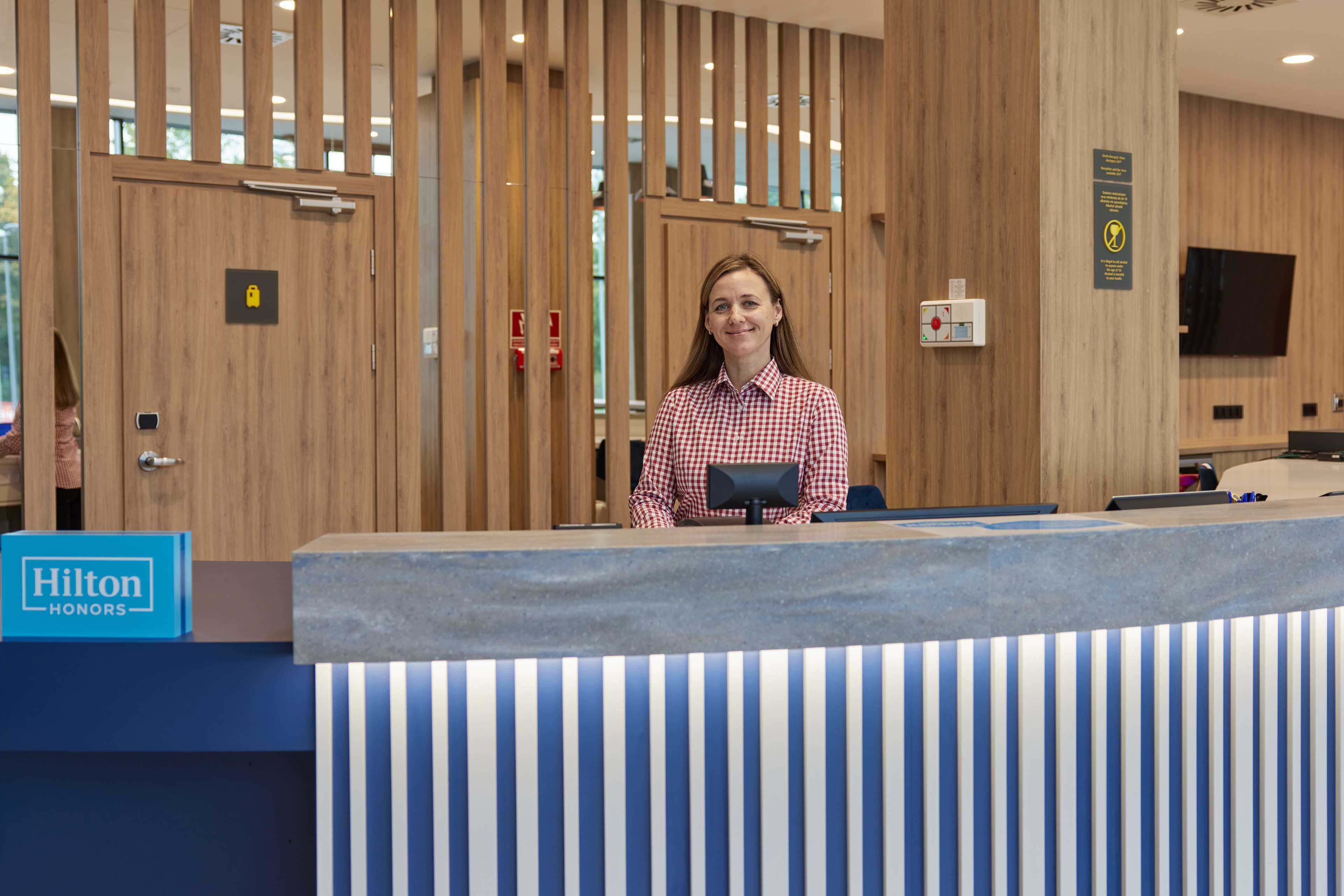 A reception desk at a Hilton hotel with a check-in counter, a sign indicating the hotel name, and a fire alarm on the wall.