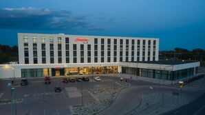 Un hotel moderno con un llamativo letrero de "Hampton", un estacionamiento con varios coches y una entrada bien iluminada.