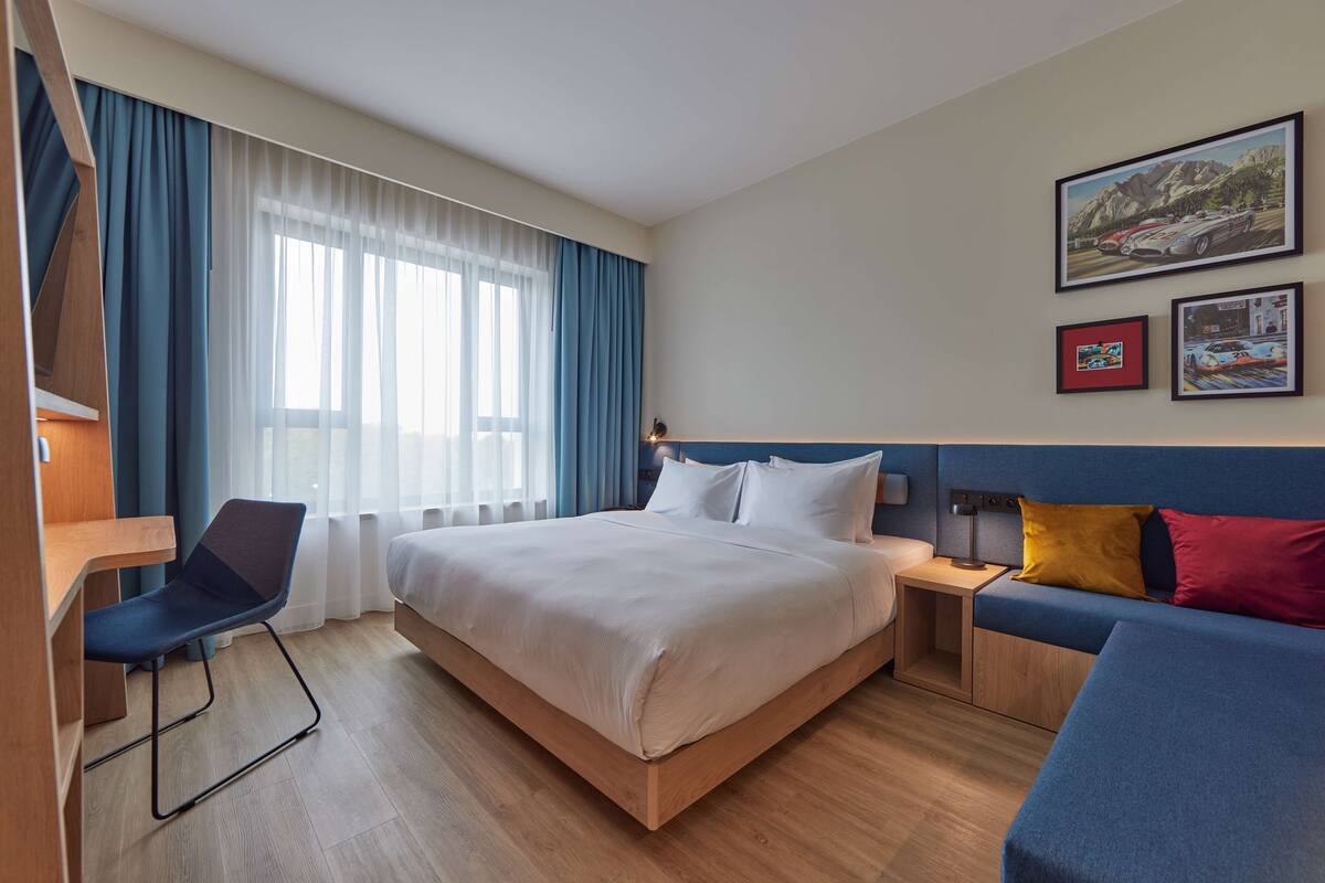 A hotel room with a bed, a desk, a chair, a sofa, and a window with curtains.