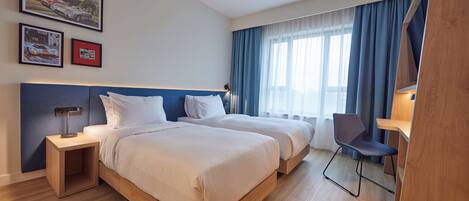 Ein Hotelzimmer mit zwei Betten, einem Schreibtisch und einem Sessel. An der Wand befinden sich Bilder in Rahmen und ein Fenster mit Vorhängen.