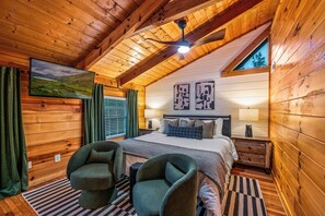 Une chambre confortable, aménagée dans une cabane en bois, avec un lit, deux fauteuils, un téléviseur et un ventilateur de plafond.