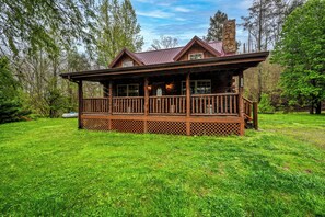 Une cabane en bois dotée d’un porche et d’une cheminée en pierre, entourée d’un paysage verdoyant et luxuriant.