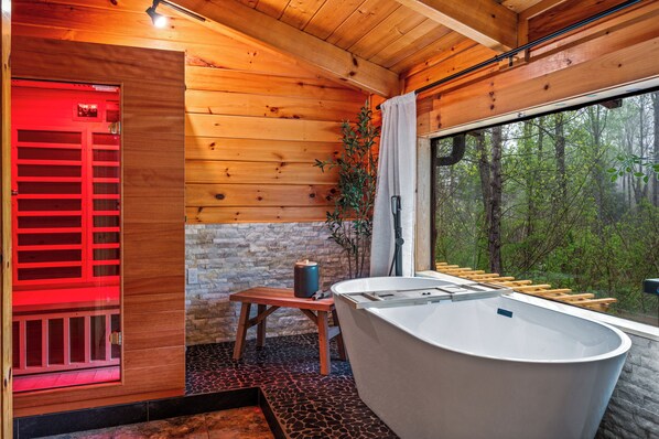 A wooden cabin interior with a large bathtub, a stone wall, and a view of a forest through a window.