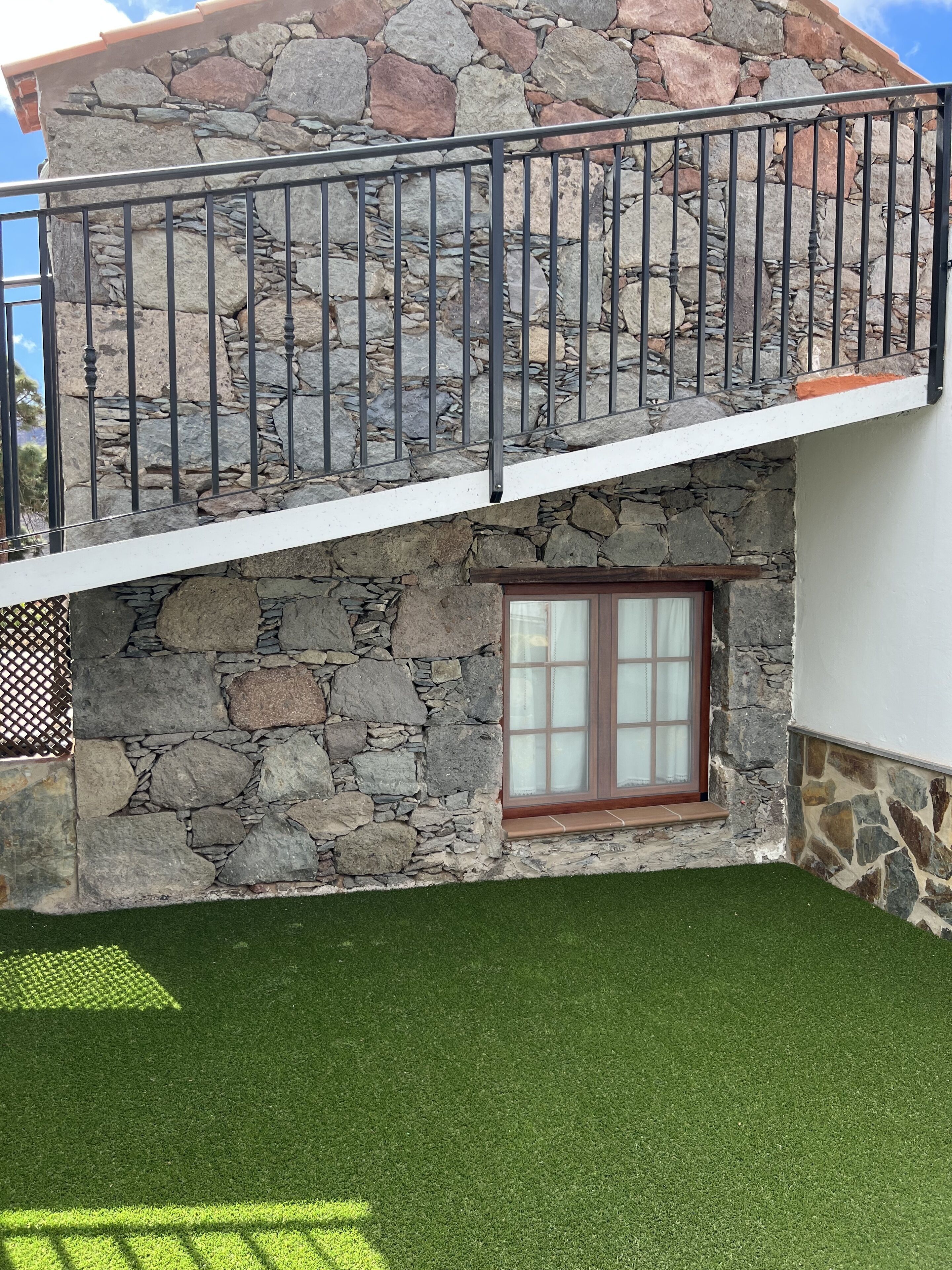 A two-story stone house with a balcony, a window with curtains, and a well-maintained lawn.