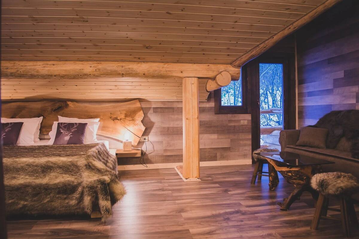 A cozy cabin bedroom with a wooden bed, a nightstand, a window with a view of trees, and a rustic wooden chair.