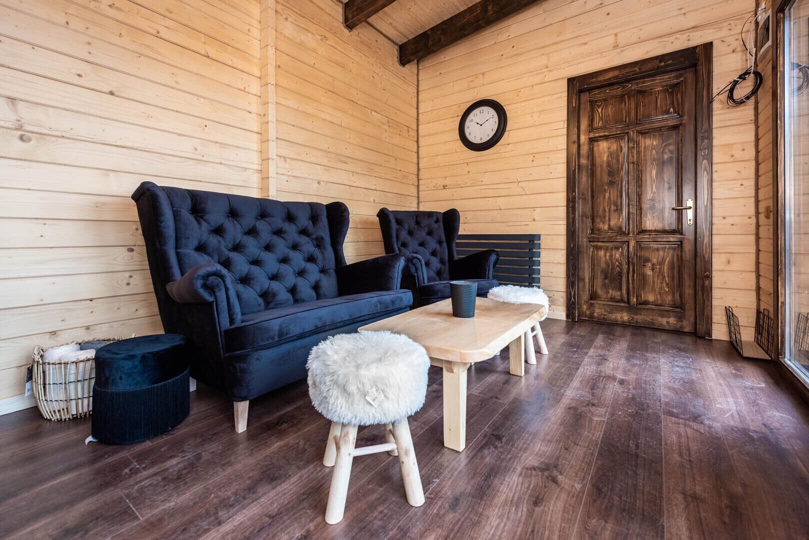 Une pièce chaleureuse avec un canapé bleu foncé, deux fauteuils, une table en bois et un petit tabouret moelleux. On peut voir une porte en bois et une horloge murale.