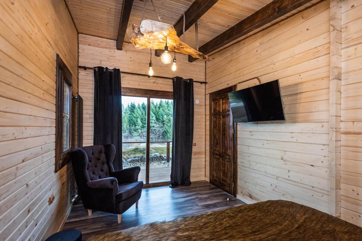 Une pièce chaleureuse avec une grande fenêtre, un fauteuil confortable, un plancher en bois et un téléviseur à écran plat fixé au mur.