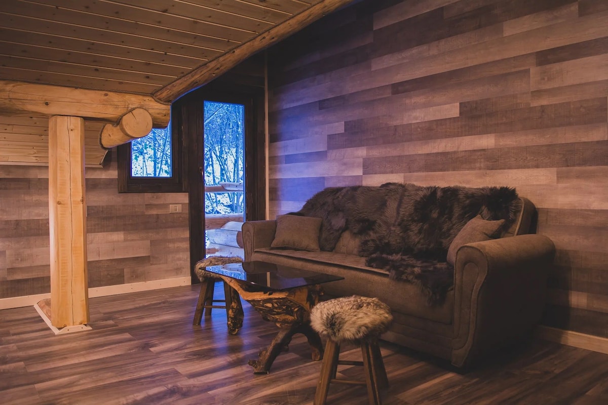 A cozy room with a wooden log wall, a sofa, a wooden coffee table, and a window with a view of a snowy landscape.