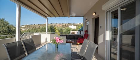 Ein Balkon mit einem Glastisch, Stühlen und einem Blumenstrauß. Im Hintergrund sind ein roter Sessel und eine Couch zu sehen.