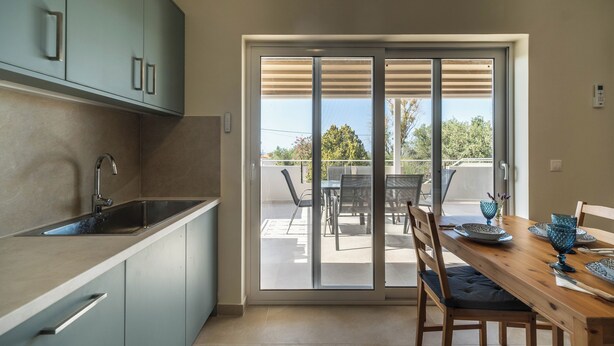 A modern kitchen with a built-in sink and cabinets, a dining area with a wooden table and chairs, and a sliding glass door leading to an outdoor patio. - 2 Bed House - Sleeps 5 - Garden - Parking - Wifi (Apokoronas)