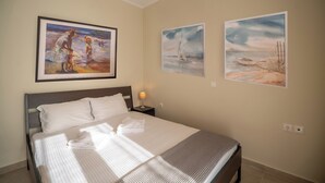 A neatly made bed with white linens and a folded towel, a nightstand with a lamp, and three paintings on the wall depicting serene beach scenes. - 2 Bed House - Sleeps 5 - Garden - Parking - Wifi (Apokoronas)